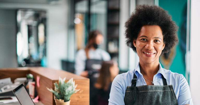 happy african american hairstylist standing with arms crossed at hair salon and looking at camera.